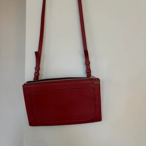 Mörkröd axelväska i skinn från Filippa K - Snygg röd axelväska från Filippa K i äkta skinn med justerbar axelrem och guldfärgade metalldetaljer. Väskan har en stor ytterficka framtill och dragkedja upptill. Perfekt storlek för det viktigaste och stilren design som sticker ut. Kommer med original dust bag. 