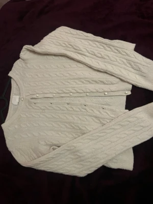 Vit kabelstickad kofta med pärlknappar - Supersöt vit kabelstickad kofta med pärlknappar framtill. Koftan har rund halsringning, långa ärmar och ribbade muddar. Perfekt för lager-på-lager och ger en klassisk preppy vibe.