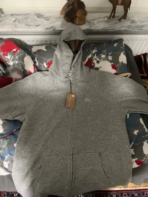 Burrbery kofta  - En hoodie från Burberry i ljusgrått. Den har dragkedja framtill, huva med snörning och stora fickor. Insidan av huvan har ett klassiskt Burberry-mönster. Perfekt för dig som gillar stilrena och bekväma plagg med exklusiv känsla. 