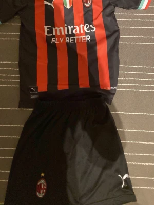 AC Milan matchtröja Puma svart/röd - Snygg AC Milan matchtröja från Puma i svart och rött med klubbmärke, sponsortryck och italienska flaggan på bröstet. Korta ärmar med detaljer i grönt, vitt och rött. Perfekt för fotbollsfans som vill representera sitt lag.