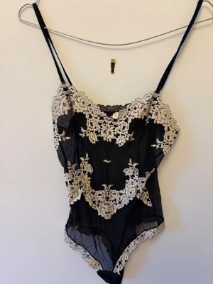 Intimissimi body - Säljer en svart mesh-body med tunna axelband och vackra vita spetsbroderier. Bodyn har genomskinligt material och dekorativa spetsdetaljer längs kanterna. Perfekt för dig som gillar en elegant och feminin stil.