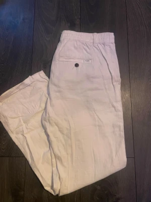 Beiga linnebyxor med dragsko - Säljer ett par beiga linnebyxor med avslappnad passform och dragsko i midjan. Byxorna har resår i midjan, sidofickor och en bakficka med knapp. Perfekta för en chill och stilren look. Materialet är luftigt linne som känns skönt mot huden.