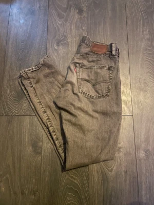 Grå Levi's jeans med rak passform - Säljer ett par grå jeans från Levi's med klassisk rak passform och fem fickor. Jeansen har en brun läderpatch bak i midjan och den ikoniska röda Levi's-taggen på bakfickan. Tillverkade i slitstarkt denimtyg, perfekta för en avslappnad stil.