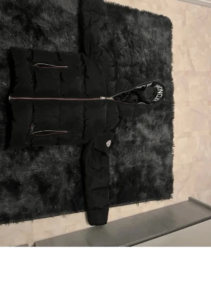 Svart dunjacka från Moncler - Snygg svart dunjacka från Moncler med stor huva och tydlig Moncler-logga på huvkanten. Jackan har två dragkedjeförsedda fickor fram och en Moncler-patch på ärmen. Puffig modell som ger en cool och modern look. Perfekt för kalla vinterdagar.