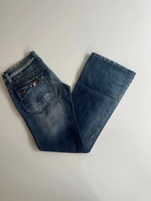 Blå bootcut replay jeans  - Säljer ett par blå bootcut jeans med slitningar och snygg tvätt. Bakfickan har broderade detaljer och färgglada stenar som ger en unik touch. Jeansen har klassisk femficksmodell och rutigt innerfoder i midjan. WV 474B färg: 032. Däremot trasig dragkedja som går att byta eller fixa lätt. 