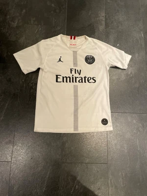 Vit PSG matchtröja Neymar Jr - Säljer en vit PSG matchtröja med Neymar Jr och nummer 10 på ryggen. Tröjan har Fly Emirates-tryck på framsidan, PSG-logga och Jordan-logga. Materialet är lätt och andas, perfekt för fotboll eller träning. Snygga röda detaljer vid kragen.