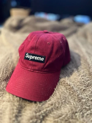 Vinröd Supreme keps - Säljer en vinröd keps från Supreme med vit broderad logga baktill och justerbart spänne i mässing. Insidan har Supreme-mönstrat band och kepsen är tillverkad i bomull. Perfekt för dig som vill ha en streetwear-vibe. 79 kr frakt