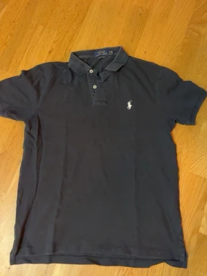 Mörkblå pikétröja från Polo Ralph Lauren - Klassisk mörkblå pikétröja från Polo Ralph Lauren med vit broderad logga på bröstet. Tröjan har korta ärmar, krage och knappar framtill. Perfekt till jeans eller shorts för en clean och stilren look.