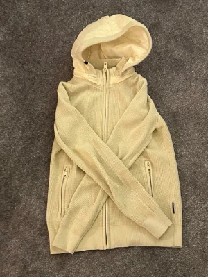 Beige hoodie med dragkedja och huva - Säljer en beige hoodie från indicode jeans med dragkedja framtill och två fickor med dragkedja. Hoodien har en quiltad, fodrad huva och är gjord i ett stickat material som känns både mjukt och stilrent. Perfekt för dig som gillar en clean och avslappnad look.