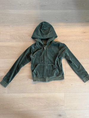  Juicy Couture - Grön velour hoodie - Mjuk grön hoodie i velour från Juicy Couture med dragkedja framtill, fickor och broderad logga på bröstet. Klassisk huva med snören och ribbade muddar vid ärmslut och nederkant. 
