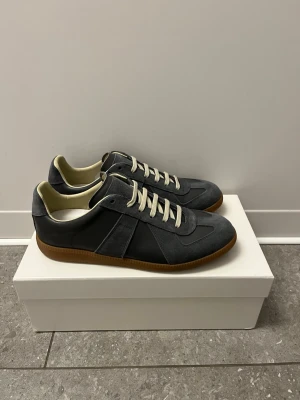Maison Margiela grå sneakers i mocka och skinn - Maison Margiela sneakers i grå mocka och skinn med vita snören och beige innersida. Snygg kontrast med gummisula i brunt och klassisk siluett. Perfekta för dig som gillar stilrena och tidlösa sneakers med premiumkänsla. Tveka inte att höra av er vid minsta fråga eller fundering. Pris går att diskuteras vid snabb affär