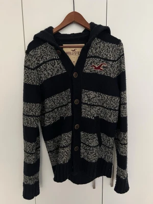 Randig kofta från Hollister - Mysig kofta från Hollister med breda svarta och gråmelerade ränder, luva och knäppning framtill. Tröjan har långa ärmar, ribbade muddar och två fickor. Hollister-logga broderad på bröstet. Perfekt för en avslappnad stil.