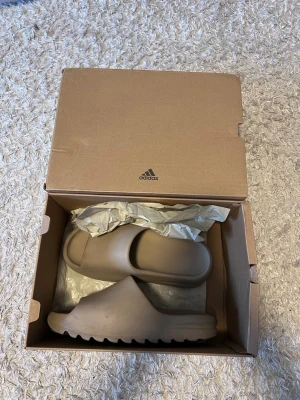 Yeezy slides  - Säljer ett par Yeezy Slide i färgen Pure från Adidas. Tofflorna har en minimalistisk design med bred rem s  och räfflad sula för extra grepp. Tillverkade i ett mjukt och flex, skönt material som passar perfekt för chill dagar sopmmar eller hemma.