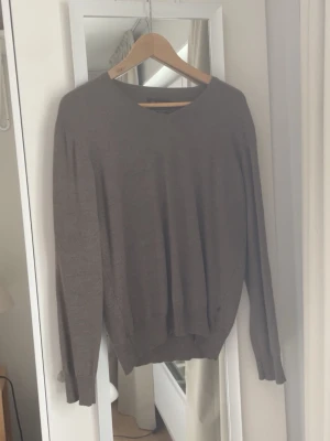 Stickad tröja – Riley - Stilren stickad tröja från Riley i en snygg brun/grå nyans. Klassisk design med v-ringning som ger ett rent och välklätt uttryck. Ett mångsidigt plagg som funkar perfekt både till vardags och mer uppklädda outfits. ✨ Passform: Bekväm och tidlös ✨ Skick: Mycket fint skick ✨ Enkel att matcha – perfekt basplagg för höst & vinter