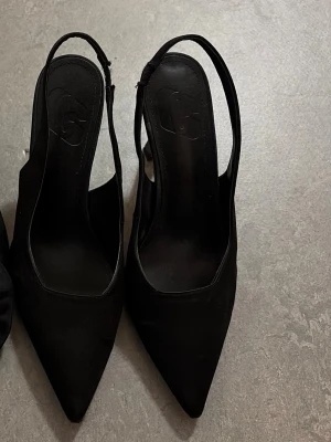 Svarta spetsiga slingback-pumps - Snygga svarta pumps med spetsig tå och slingback-rem. Skorna har en elegant klack och är tillverkade i ett tygmaterial med slät finish. Perfekta för att lyfta vilken outfit som helst med en stilren look.