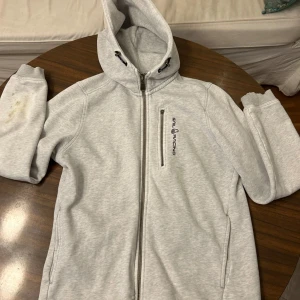 Ljusgrå hoodie från Sail Racing - Ljusgrå hoodie från Sail Racing med hel dragkedja och huva med snörning. Tröjan har en bröstficka med dragkedja och Sail Racing-logga på framsidan. Mjuk insida i fleece och långa ärmar med ribbade muddar. Perfekt för chill eller utomhus. Den har ganska många fläckar på ärmen!
