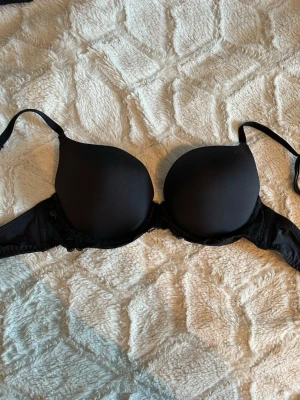 Svart push-up bh med spetsdetaljer - Snygg svart push-up bh från Hunkemöller med vadderade kupor och fina spetsdetaljer på sidorna. Justerbara axelband och hakknäppning bak. Perfekt för dig som vill ha extra lyft och en klassisk look.