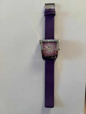 Lila fyrkantig klocka från dolce & gabbana  - Snygg klocka från dolce & gabbana med fyrkantigt silverfärgat boett och lila urtavla. Armbandet är i matchande lila syntetmaterial och har klassiskt spänne. Urtavlan har tydliga siffror och en modern look som sticker ut. Behöver nytt batteri 