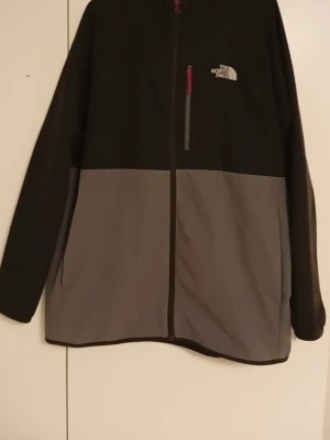 Svart och grå jacka vind/skaljacka från North Face - Säljer en svart och grå jacka från The North Face, Nypris var 1000kr,  Stl. (XL) men passar även (L) med hel dragkedja och bröstficka med röd detalj. Jackan har långärmad design och logotyp på bröstet. Perfekt för kyliga dagar och enkel att matcha med olika outfits. Du kan nå mig på 0737013526