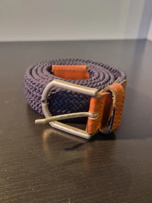 Mörkblått flätat bälte med skinn - Snyggt mörkblått flätat bälte med detaljer i brunt skinn och metallspänne. Perfekt accessoar för att lyfta din outfit med en klassisk touch. Bältet har en flexibel och justerbar design tack vare det elastiska materialet.