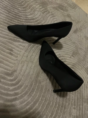Svarta klassiska klackar med smal klack - Säljer ett par svarta klackar med spetsig tå och smal klack. Skorna har en stilren och tidlös design, perfekta för dig som gillar en elegant look. Materialet ser ut att vara textil eller syntet. Passar till många olika outfits. Har aldrig använts 