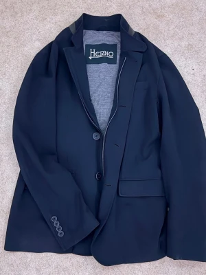  Herno jacka/kavaj - Stilren svart kavaj från Herno med klassisk passform och två knappar framtill. Kavajen har grått innerfoder och detaljer vid axlarna. Perfekt för en dressad look och tillverkad i ett mjukt material som känns bekvämt hela dagen.