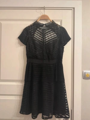 Svart spetsklänning By Malina - Supersnygg svart kortklänning med spetsdetaljer och hög hals. Klänningen har korta ärmar, markerad midja och transparenta partier upptill. Perfekt för dig som gillar en elegant och trendig look. Spetsen ger en lyxig känsla och den har en lätt utställd kjol.