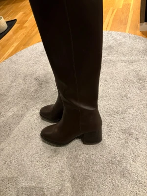 Bruna knähöga läderstövlar - Snygga bruna knähöga stövlar med rund tå och bred klack. Tillverkade i slätt läder med diskreta sömmar längs sidan. Storlek 38. Använd ca 3 ggr 