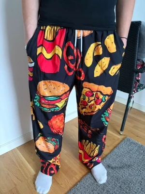 Svarta loungewear-byxor med matmotiv - Färgglada loungewear-byxor med svart bas och stora tryck av hamburgare, pizza, pommes och andra snacks. Byxorna har resår i midjan med dragsko och sidofickor. Perfekta för chill hemma och för dig som älskar roliga mönster.