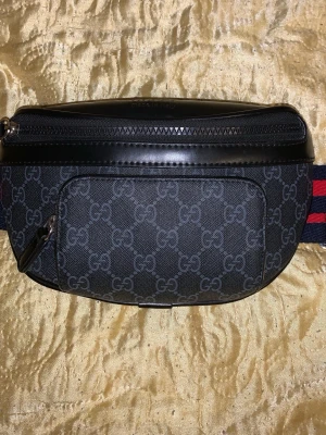 Gucci svart shoppingväska med monogram - Snygg svart shoppingväska från Gucci med klassiskt GG-monogram i grått. Väskan har detaljer i svart läder, dragkedja upptill och en bred axelrem i marinblått och rött. Perfekt för dig som vill ha en trendig och praktisk accessoar.