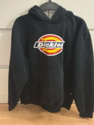 Svart Dickies hoodie med logga - Svart hoodie från Dickies med stor färgglad logga på bröstet. Klassisk modell med huva och känguruficka framtill. Tillverkad i en mjuk bomulls- och polyestermix som är skön att ha på sig. Perfekt för en avslappnad streetwear-look.
