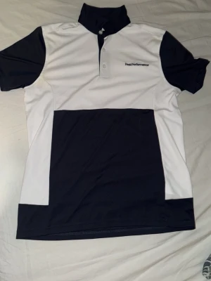Peak Performance sportpiké svart/vit - Sportig pikétröja från Peak Performance i svart och vitt. Tröjan har korta ärmar, knappar vid halsen och logga broderad på bröstet. Perfekt för träning eller golf. Tillverkad i ett lätt och funktionellt material som andas.