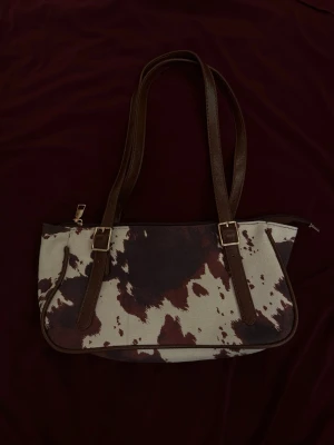 Brun och vit handväska med koskinnsmönster - Säljer en trendig handväska med koskinnsmönster i brunt och vitt. Väskan har två långa axelband i brunt konstläder och guldfärgade spännen. Perfekt för dig som vill sticka ut med en unik accessoar!