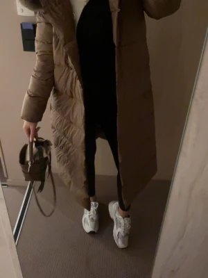 Lång brun dunjacka med pälskrage - Snygg lång brun dunjacka med bälte i midjan och stor pälskantad huva. Jackan har quiltad design och stängs med band i midjan och knappar. Perfekt för kalla vinterdagar och ger en riktigt trendig look.