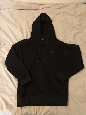 Svart hoodie från Polo Ralph Lauren - Säljer en svart hoodie från Polo Ralph Lauren med dragkedja och huva. Klassisk röd logga broderad på bröstet. Hoodien har ribbade muddar och dragsko i huvan. Perfekt för en chill och stilren look.