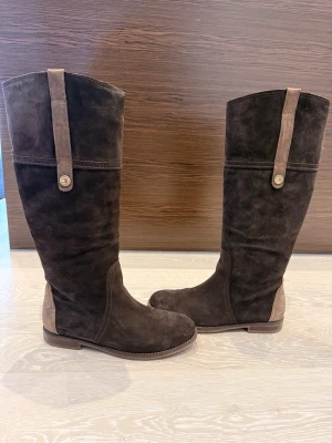 Bruna mockastövlar Tommy Hilfiger - Snygga bruna stövlar i mocka från Tommy Hilfiger med dekorativa remmar och knappdetaljer. Stövlarna har låg klack och kontrasterande beige detaljer vid häl och skaft. Perfekta för dig som vill ha en stilren och klassisk look.