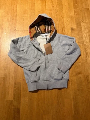 Burberry kofta - Säljer en grå zip-hoodie från Burberry med klassiskt rutigt foder i huvan. Hoodien har dragkedja framtill, två fickor och ribbade muddar vid ärmar och nederkant. Perfekt för en avslappnad och stilren look. Den passar för de som har size xs-s