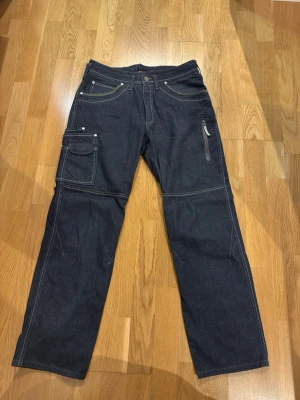 Vintage mörkblå cargobyxor med fickor - Snygga vintage  mörkblå cargobyxor i denim med flera praktiska fickor, bland annat en med dragkedja på benet och en klassisk cargoficka. Byxorna har raka ben och kontrastsömmar som ger en cool look. Perfekta för dig som gillar streetwear och funktionella plagg.