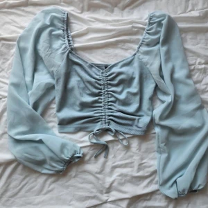 Ljusblå croppad topp med snörning - Trendig ljusblå croppad topp från Bershka med långa puffärmar i transparent tyg och snygg snörning framtill. Toppen har ribbad struktur på livet och en feminin v-ringning. Perfekt för dig som vill sticka ut med en cool och söt stil.