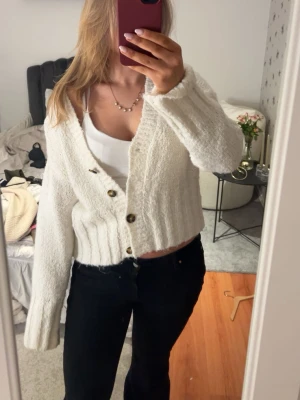 Vit stickad kofta med knappar - Mysig vit stickad kofta med ribbad midja och stora bruna knappar framtill. Koftan har lång ärm och en något croppad passform som ger en trendig look. Perfekt att slänga över ett linne eller t-shirt för en avslappnad stil.