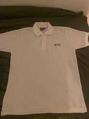 Vit pikétröja från BOSS - Klassisk vit pikétröja från BOSS med kort ärm och krage. Tröjan har knappar framtill och en diskret BOSS-logga på bröstet. Perfekt för en clean och stilren look. Använd 1 gång annars helt ny och fint skick.