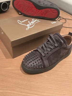 Gråa Christian Louboutin - Snygga gråa sneakers från Christian Louboutin i mocka med ikoniska nitar på tån och röd sula. Allt följer med dvs dustbag, kvitto, kort och två extra spikar. Pris är inte ristat i sten, hör av er vid funderingar!💯💸