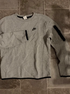 Nike tech sweatshirt  - Snygg grå sweatshirt från Nike med svart logga på bröstet. Tröjan har svarta detaljer vid dragkedjor på bröstet och ena ärmen, samt rund halsringning och ribbade muddar. Perfekt för en sportig och avslappnad stil.