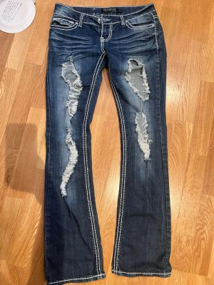 Mörkblå slitna bootcut jeans PREMIERE - Säljer ett par mörkblå bootcut jeans från PREMIERE med kraftiga slitningar och hål framtill. Jeansen har kontrastsömmar och bakfickor med broderade detaljer och knappar. Perfekta för en edgy och trendig look. 