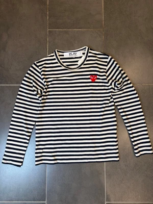 Randig långärmad topp Comme des Garçons - Snygg långärmad topp från Comme des Garçons PLAY med svartvita ränder och det ikoniska röda hjärtat broderat på bröstet. Tillverkad i mjuk bomull och har en klassisk rund halsringning. Perfekt för dig som gillar streetstyle och vill ha något tidlöst.