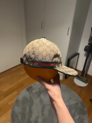 Beige Gucci keps - Säljer en beige keps från Gucci med det klassiska GG-monogrammet över hela kepsen. Kepsen har en böjd skärm och justerbart band bak med den ikoniska gröna och röda Gucci-remsan. Perfekt för dig som vill ha en stilren och lyxig accessoar.