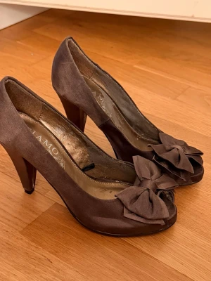 Bruna pumps med rosett från Tiamo - Snygga bruna pumps från Tiamo med peep-toe och stora dekorativa rosetter framtill. Tecken på andväning men en ny sula för uppfräschning!