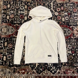 Vit hoodie från Carlheim - Snygg vit hoodie från Carlheim med klassisk huva och stor magficka. Tillverkad i mjukt material och har en diskret svart logotyp-patch på fickan. Perfekt för en clean och stilren look. Går gärna ner i pris vid snabb affär