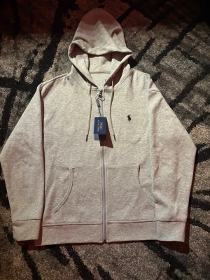 Grå zip hoodie från Polo Ralph Lauren - Snygg gråmelerad hoodie med dragkedja från Polo Ralph Lauren. Tröjan har huva med dragsko, två fickor framtill och den klassiska loggan broderad på bröstet. Perfekt för en chill och avslappnad stil. Den är M men den passar bättre S