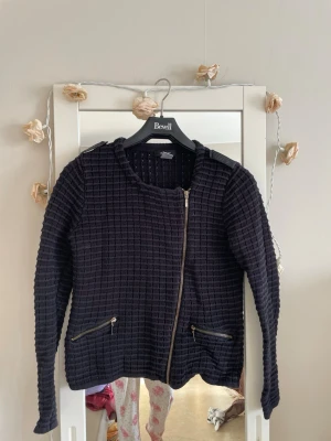 Svarts cardiganjacka med dragkedja - Svart cardiganjacka från edition med rutmönstrad struktur, snedställd dragkedja framtill och två fickor med dragkedjor. Långärmad och rund halsringning. Perfekt för en trendig och avslappnad look.